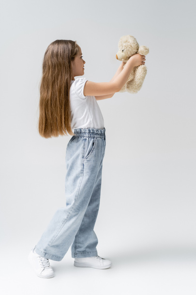 Девочка в джинсах в полный рост с плюшевым медведем на сером - Фото, изображение