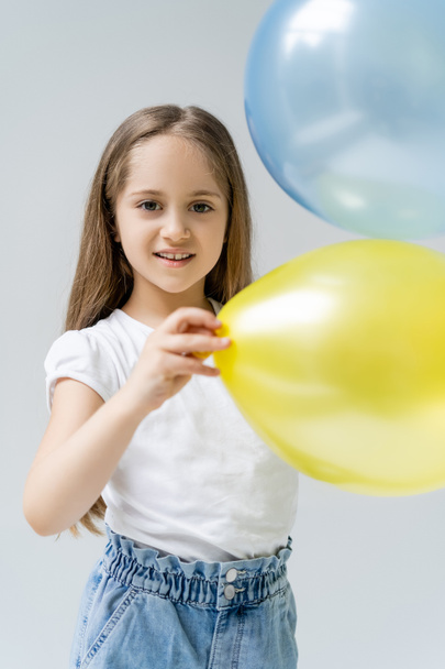zufriedenes Mädchen blickt in die Kamera in der Nähe verschwommener blauer und gelber Luftballons isoliert auf grau - Foto, Bild