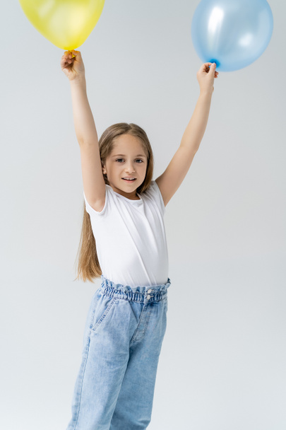 smiling girl in white t-shirt holding blue and yellow balloons in raised hands isolated on grey - Fotografie, Obrázek