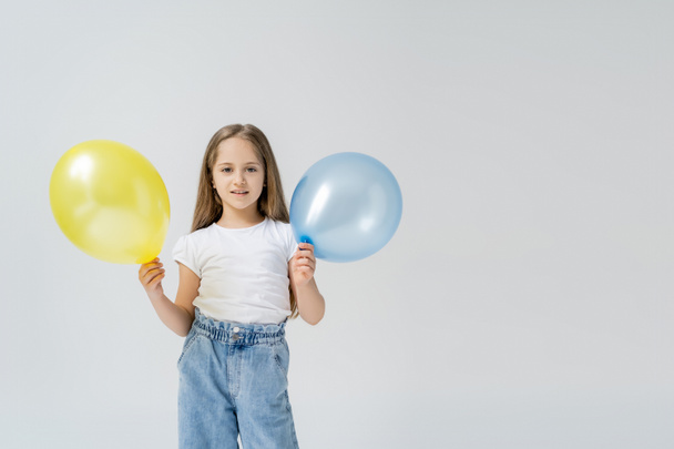 happy girl with blue and yellow balloons looking at camera isolated on grey - Zdjęcie, obraz