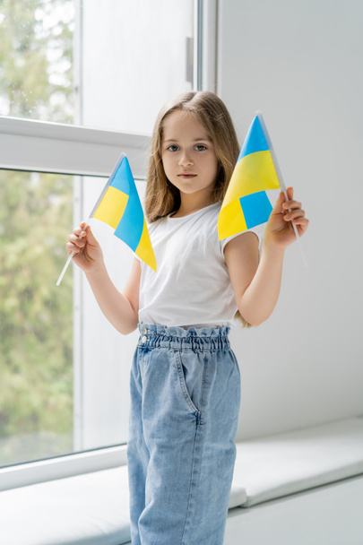 fille patriotique avec de petits drapeaux ukrainiens debout près de la fenêtre et regardant la caméra - Photo, image
