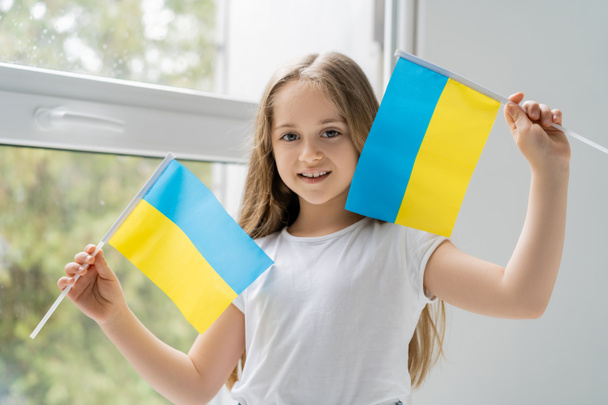 meisje met kleine Oekraïense vlaggen glimlachen in de buurt venster thuis - Foto, afbeelding