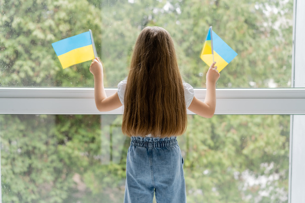 vista trasera de la chica con el pelo largo de pie con pequeñas banderas ucranianas cerca de la ventana - Foto, Imagen