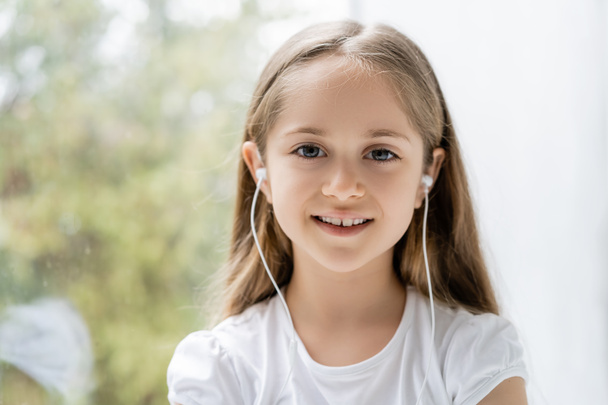 vrolijk meisje in bedraad oortelefoons glimlachen op camera in de buurt wazig venster - Foto, afbeelding
