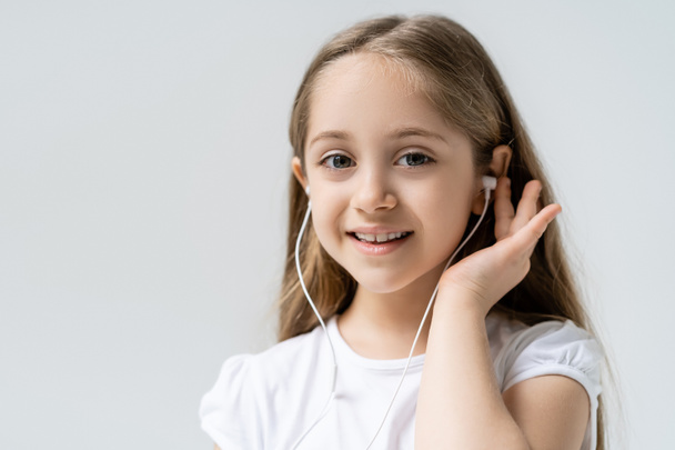 χαρούμενο κορίτσι σε ενσύρματα ακουστικά κοιτάζοντας κάμερα απομονωμένη σε γκρι - Φωτογραφία, εικόνα