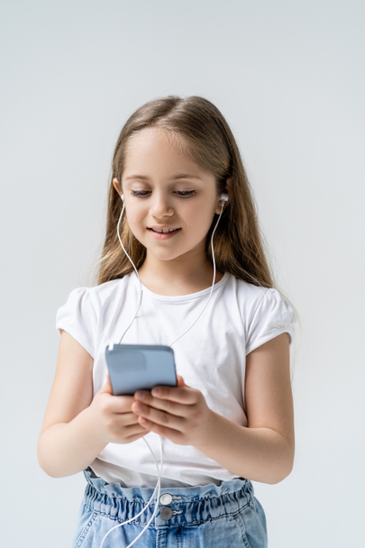 χαμογελαστό κορίτσι με ακουστικά που χρησιμοποιεί κινητό τηλέφωνο απομονωμένο σε γκρι - Φωτογραφία, εικόνα