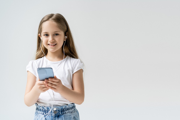 szczęśliwe dziecko ze słuchawkami i telefonem komórkowym patrząc na kamerę odizolowaną na szaro - Zdjęcie, obraz