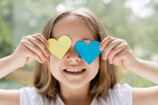 alegre chica ucraniana oscureciendo la cara con corazones de papel azul y amarillo - Foto, imagen