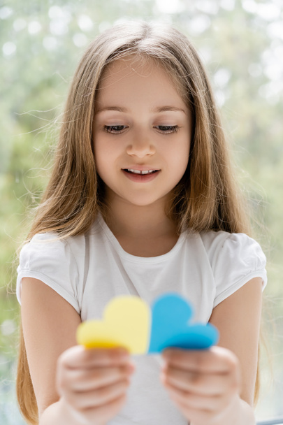 χαμογελαστό πατριωτικό κορίτσι με μακριά μαλλιά που κρατά θολές μπλε και κίτρινες χάρτινες καρδιές - Φωτογραφία, εικόνα
