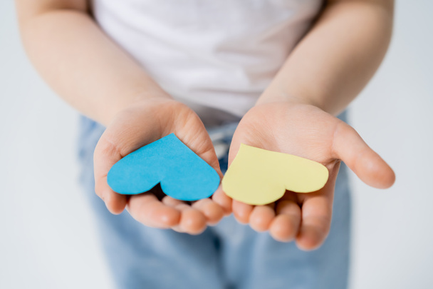 cropped view of blurred kid holding blue and yellow paper hearts in open palms isolated on grey - Photo, Image