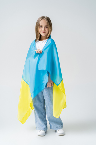 full length view of positive girl covered with ukrainian flag on grey - Photo, Image