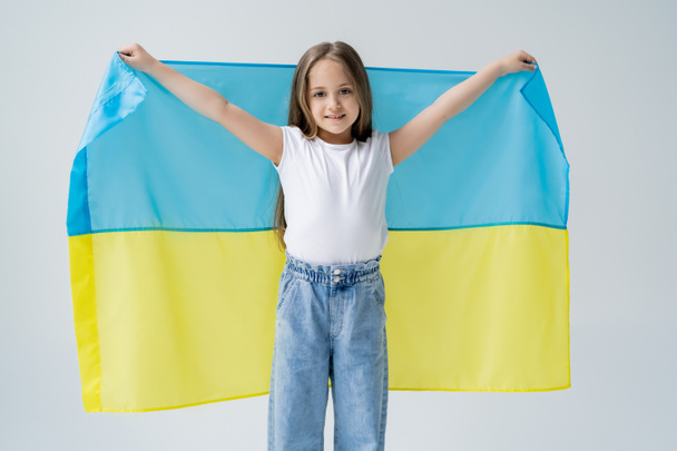 Beyaz tişörtlü ve kot pantolonlu mutlu kız gri üzerinde Ukrayna bayrağı taşıyor. - Fotoğraf, Görsel