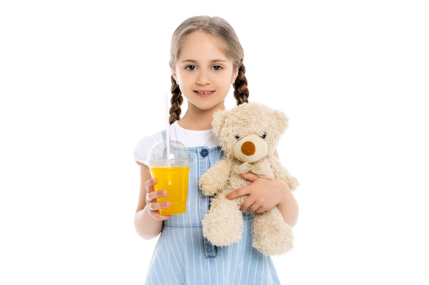chica feliz con jugo de naranja y juguete suave mirando a la cámara aislada en blanco - Foto, Imagen