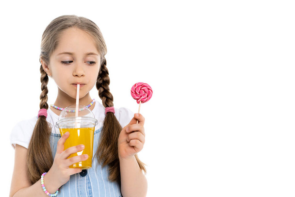 girl in colorful beads drinking orange juice and looking at lollipop isolated on white - Фото, изображение