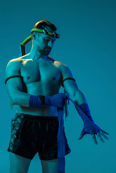 Half-length portrait of muscled man, thai boxer in sports uniform posing isolated on blue studio background in neon. Sport, muay thai, competition, fight club concept - Φωτογραφία, εικόνα