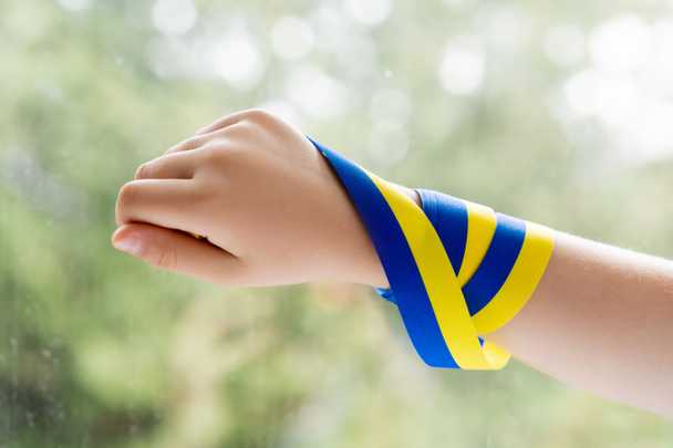cropped view of kid with blue and yellow ribbon on hand near blurred window - Fotoğraf, Görsel