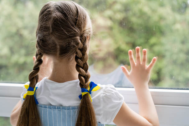 家の窓の近くに立っている橋の上に青と黄色のリボンを持つ少女の背中の景色 - 写真・画像