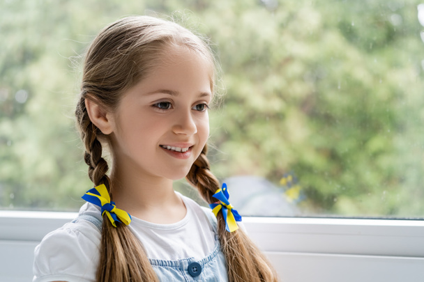 chica patriótica con cintas azules y amarillas en trenzas sonriendo cerca de ventana borrosa - Foto, Imagen