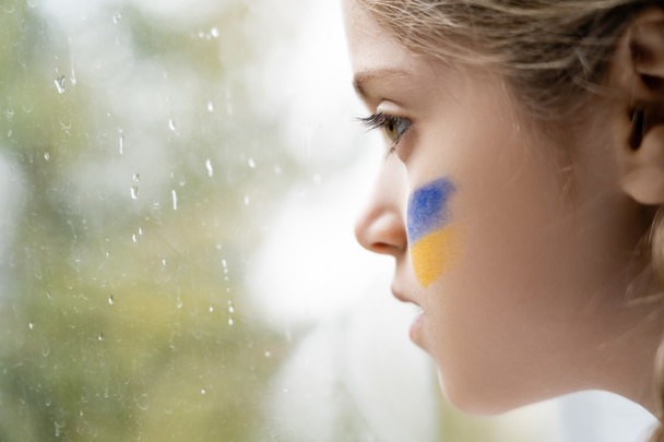 close up profile of girl with ukrainian flag painted on face near wet window glass - Photo, Image