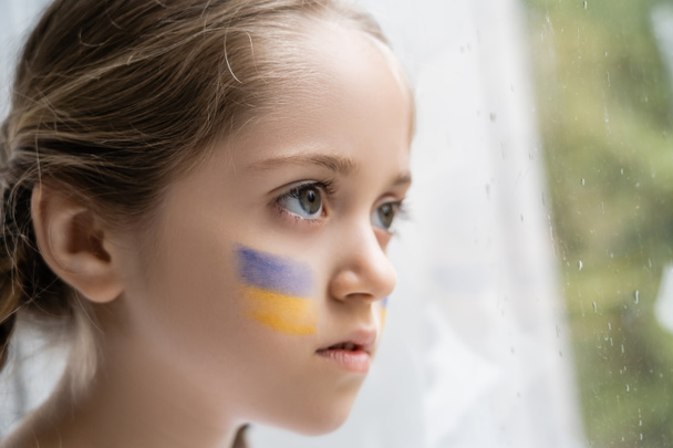 portrait of girl with ukrainian flag on face looking away through window with raindrops - Photo, Image