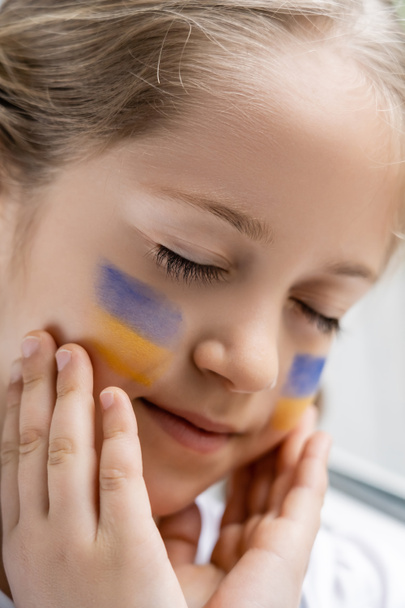 Nahaufnahme Porträt eines Mädchens mit geschlossenen Augen, das Gesicht mit bemalten ukrainischen Flaggen berührt - Foto, Bild