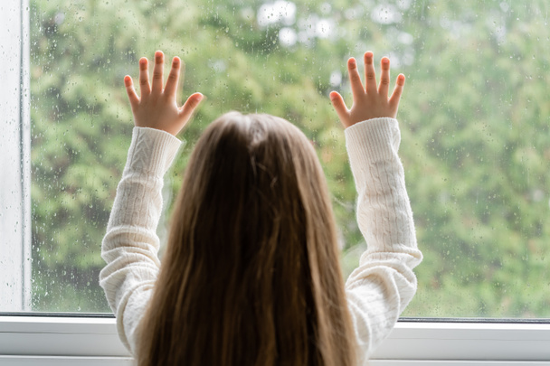 Pencerenin yanında duran ve yağmur damlalarıyla cama dokunan kızın arka plan görüntüsü - Fotoğraf, Görsel
