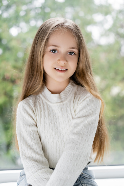 pozitivní dívka s dlouhými vlasy s úsměvem na kameru v blízkosti rozmazané okno - Fotografie, Obrázek