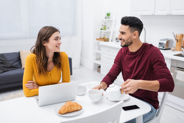 Happy multiethnic couple talking near devices and breakfast in kitchen  - Zdjęcie, obraz