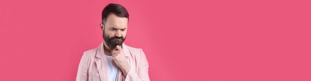 Knappe man met een baard in een roze jasje denkt over een geïsoleerde rode achtergrond. - Foto, afbeelding