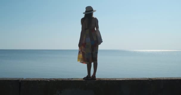 Młoda kobieta w kapeluszu stoi na betonowym pomoście i patrzy w oddali na spokojne morze. Dzień, spokojne, jasne słońce na zenicie, bezchmurne niebo. - Materiał filmowy, wideo