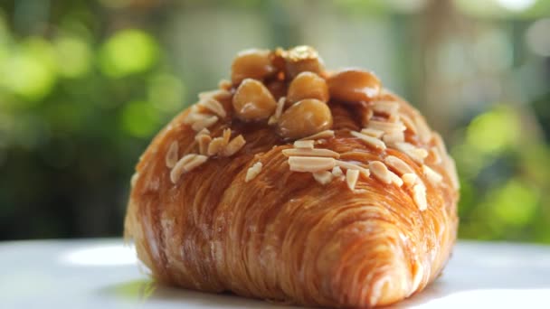 Delicious homemade croissant topped with edible gold leaf schabin flakes on macadamia, almond slivered and caramel slowly rotating  on blurred bokeh nature background. Close-up shot of high fat pastry food and dessert. - Footage, Video