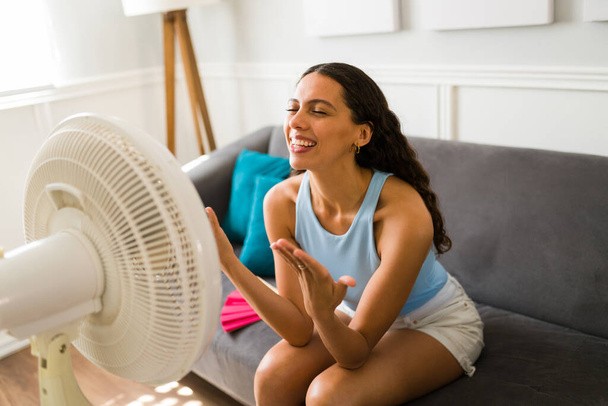 Schöne lateinische Frau ist aufgeregt, nachdem sie den elektrischen Ventilator eingeschaltet hat und die Luft wegen des warmen Wetters genießt - Foto, Bild