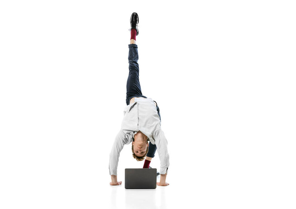 Creative portrait of young flexible man doing backbend and typing on laptop isolated over white studio background. Business in modern life concept - Fotó, kép