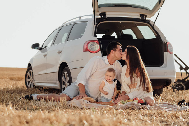 Young Family with Toddler Child Enjoying Picnic Outside the City, Mom and Dad Kissing, They are Dressed Alike with Their Daughter, Weekend Road Trip with Car - Photo, image