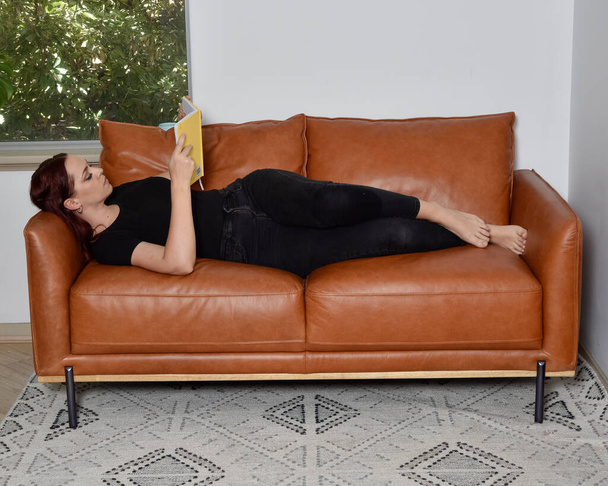 pełnowymiarowy portret rudej dziewczyny w ubraniu casual, relaks na wygodnej skórzanej kanapie w salonie ustawienie domu. - Zdjęcie, obraz