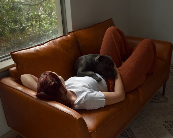 retrato de longitud completa o chica de pelo rojo con ropa casual, relajarse en el cómodo sofá de cuero en el entorno de la casa sala de estar. - Foto, imagen