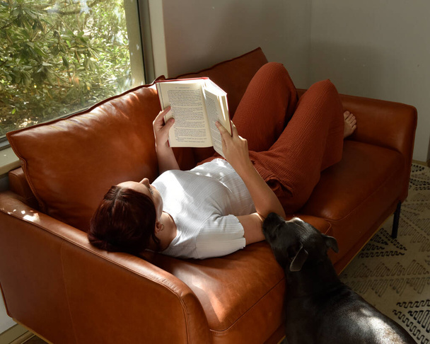 retrato de longitud completa o chica de pelo rojo con ropa casual, relajarse en el cómodo sofá de cuero en el entorno de la casa sala de estar. - Foto, Imagen