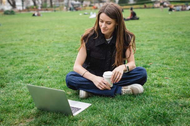 cheerful young woman holding paper cup and looking at laptop while sitting on lawn - Foto, imagen