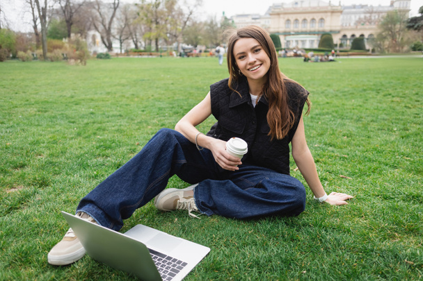 smiling young woman holding paper cup near laptop while sitting on lawn - 写真・画像