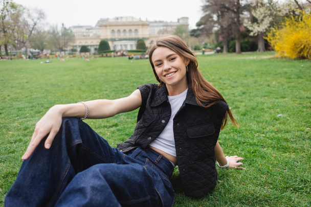fröhliche junge Frau in ärmelloser Jacke auf Rasen sitzend  - Foto, Bild