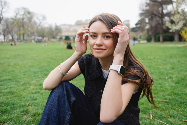 usmívající se mladá žena v bundě bez rukávů upravující vlasy při sezení na trávníku  - Fotografie, Obrázek