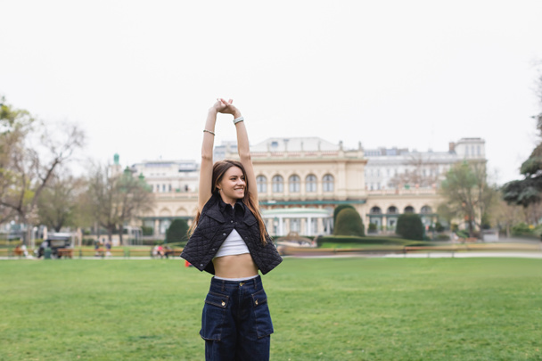 jonge vrolijke vrouw in mouwloze jas stretching in groen park  - Foto, afbeelding