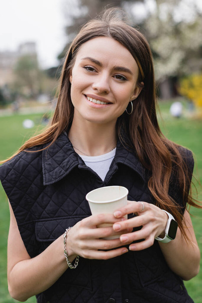young cheerful woman in sleeveless jacket holding coffee to go in green park  - Fotoğraf, Görsel