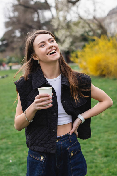 young joyful woman in sleeveless jacket holding coffee to go in green park  - Fotoğraf, Görsel
