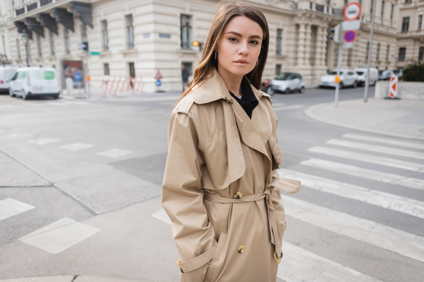 young woman in stylish trench coat standing with hands in pockets on street of european city - Photo, Image