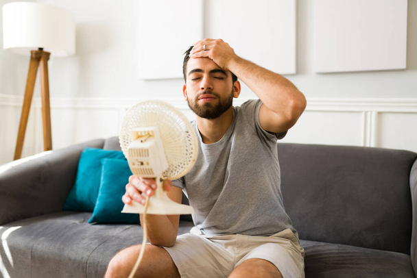 Heat stroke. Tired hispanic with his hand on the forehead feeling hot and sweating in front of the fan at home - Фото, изображение