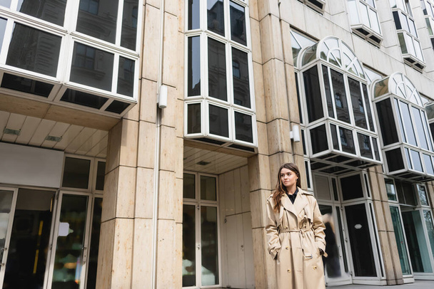 young woman in stylish trench coat standing with hands in pockets near building - Photo, Image