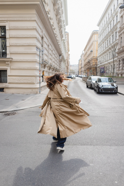 young woman in trench coat shaking head while walking in european city  - 写真・画像