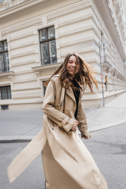 happy young woman in beige trench coat walking on european street - Photo, Image