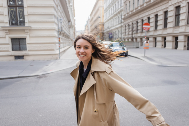 cheerful young woman in trench coat walking on street in windy day - Fotografie, Obrázek